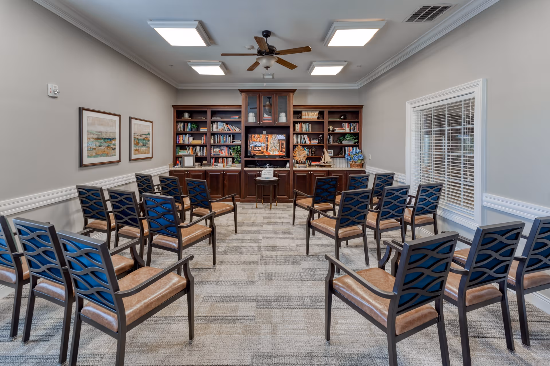 A well-lit meeting or activity room with rows of chairs facing a large wooden bookshelf and a TV screen. The room has a ceiling fan, framed artwork on the walls, and a window with blinds on the right side.