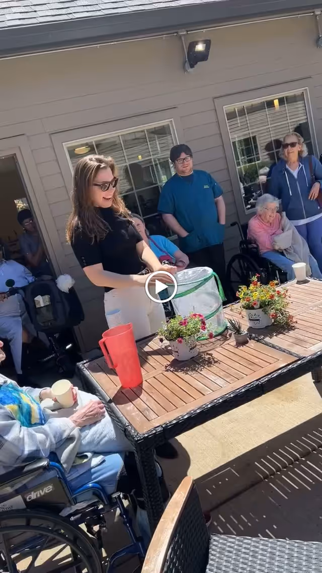 A group of elderly people and caregivers gathered outside on a patio at a senior living facility. A woman in sunglasses stands near a wooden table with potted flowers and a red pitcher, engaging with the group. Some elderly individuals are seated in wheelchairs, enjoying the sunny day.