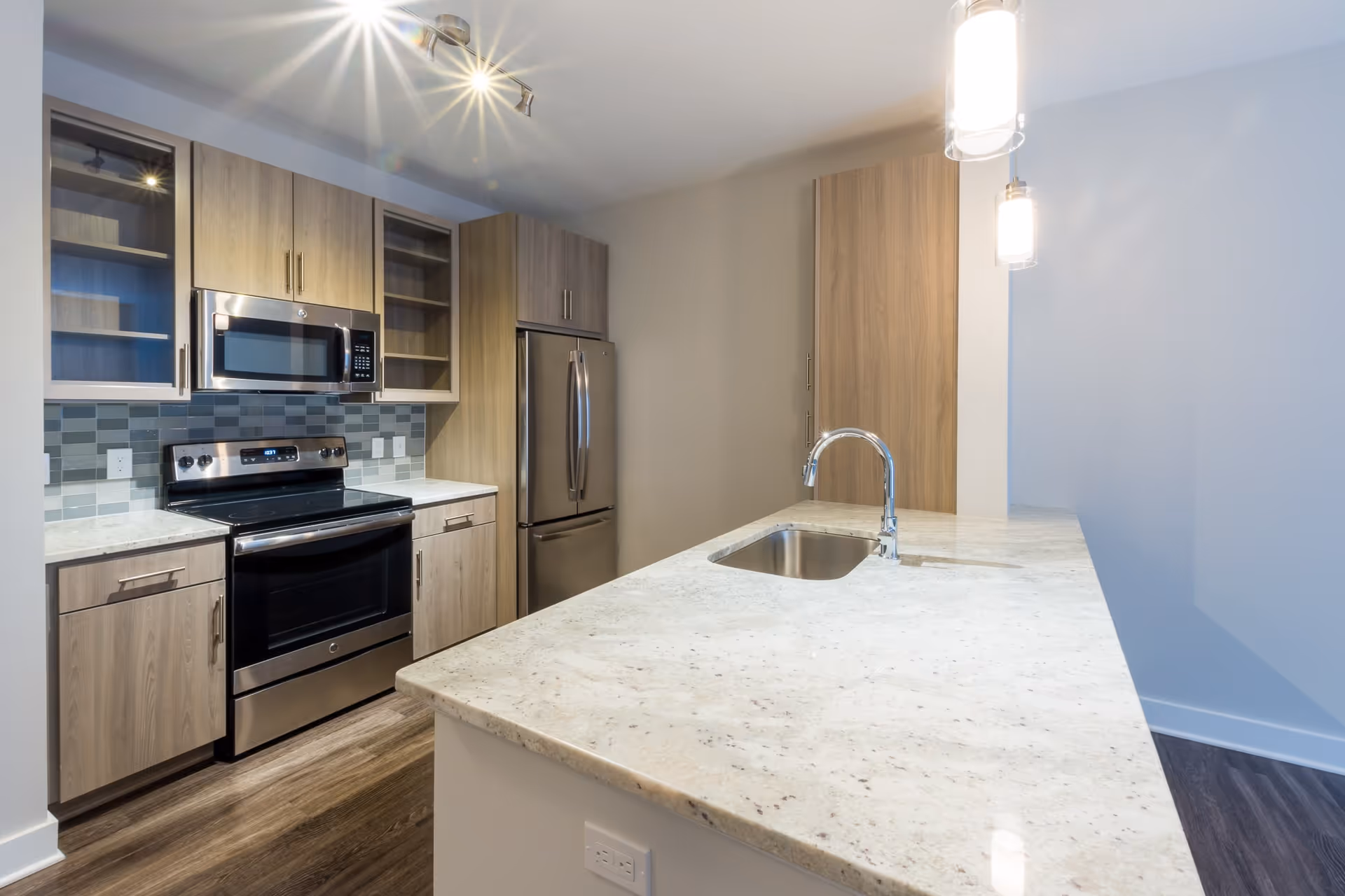 Modern kitchen with light wood cabinets, stainless steel appliances including a microwave, oven, and refrigerator, a large marble countertop island with a built-in sink, and pendant lighting above the island.