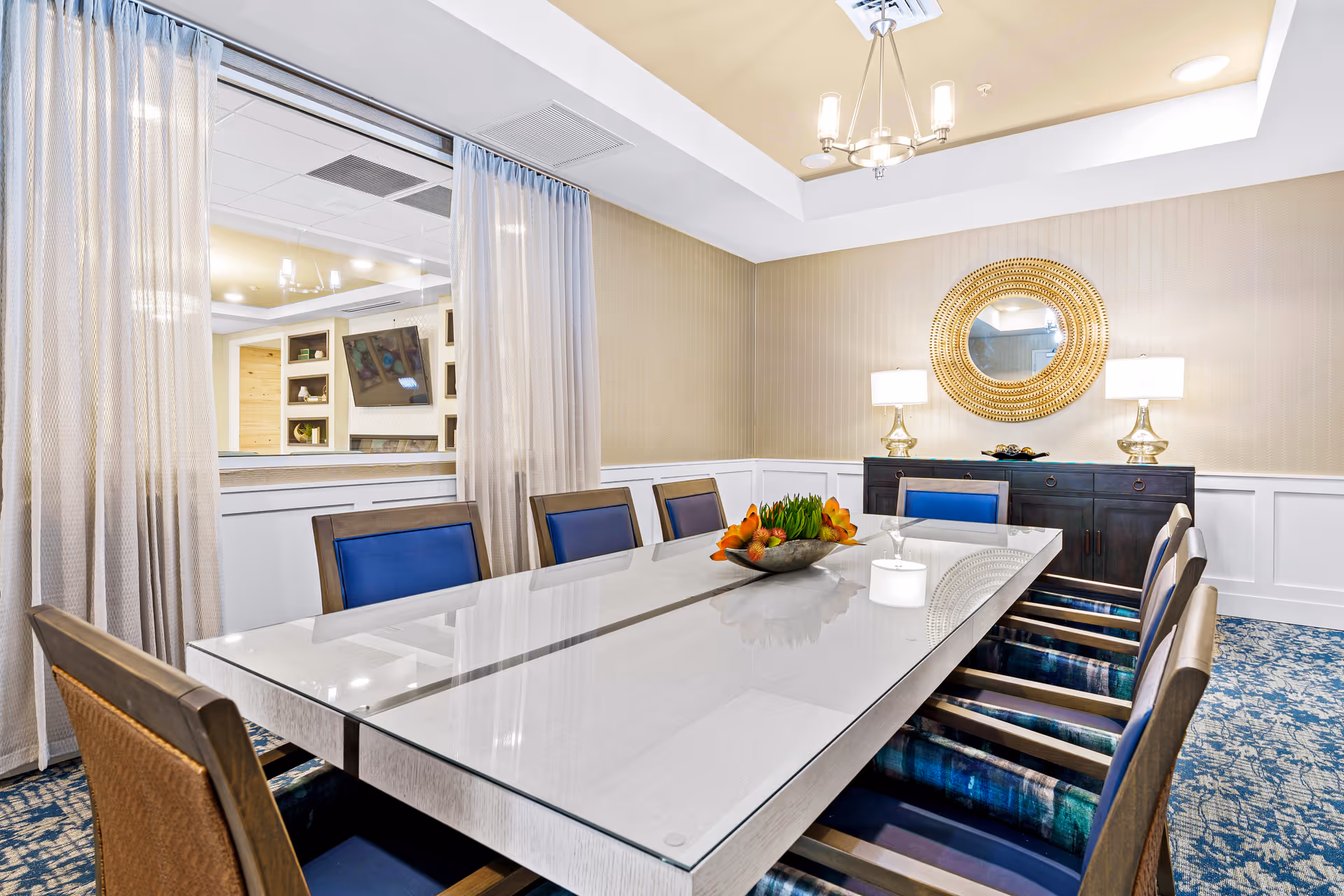 A well-lit dining room with a long rectangular glass-top table surrounded by eight chairs with blue cushions. The room features beige walls with white wainscoting, a decorative round mirror above a dark wooden sideboard with two lamps, and a centerpiece with orange flowers and greenery on the table. Sheer curtains cover a large window opening to an adjacent room with a TV and built-in shelves.