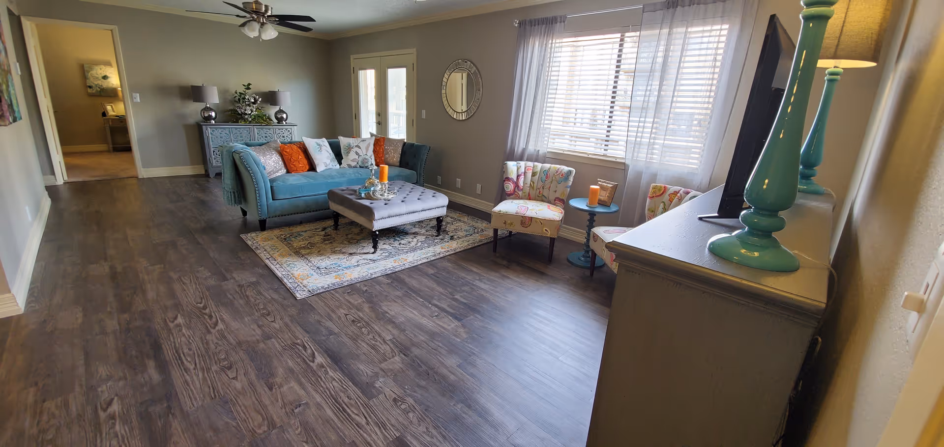 A bright and cozy living room with a teal sofa adorned with colorful pillows, a gray tufted ottoman on a patterned rug, two floral-patterned chairs with a small blue side table between them, a large window with sheer curtains, a ceiling fan, and a gray cabinet with teal lamps and a TV on top.