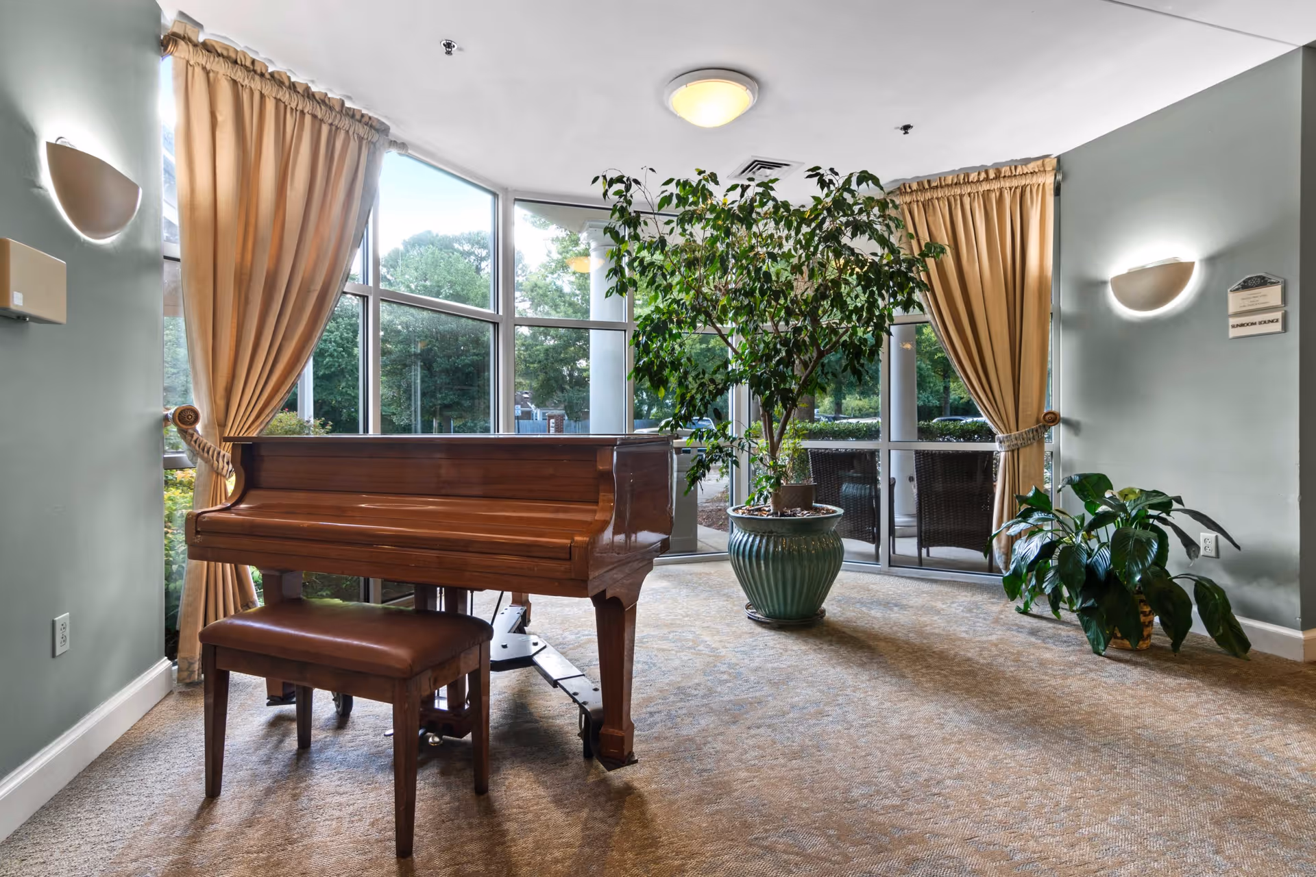 Interior common room with a wooden piano and bench by large windows, potted plants, and gold curtains.