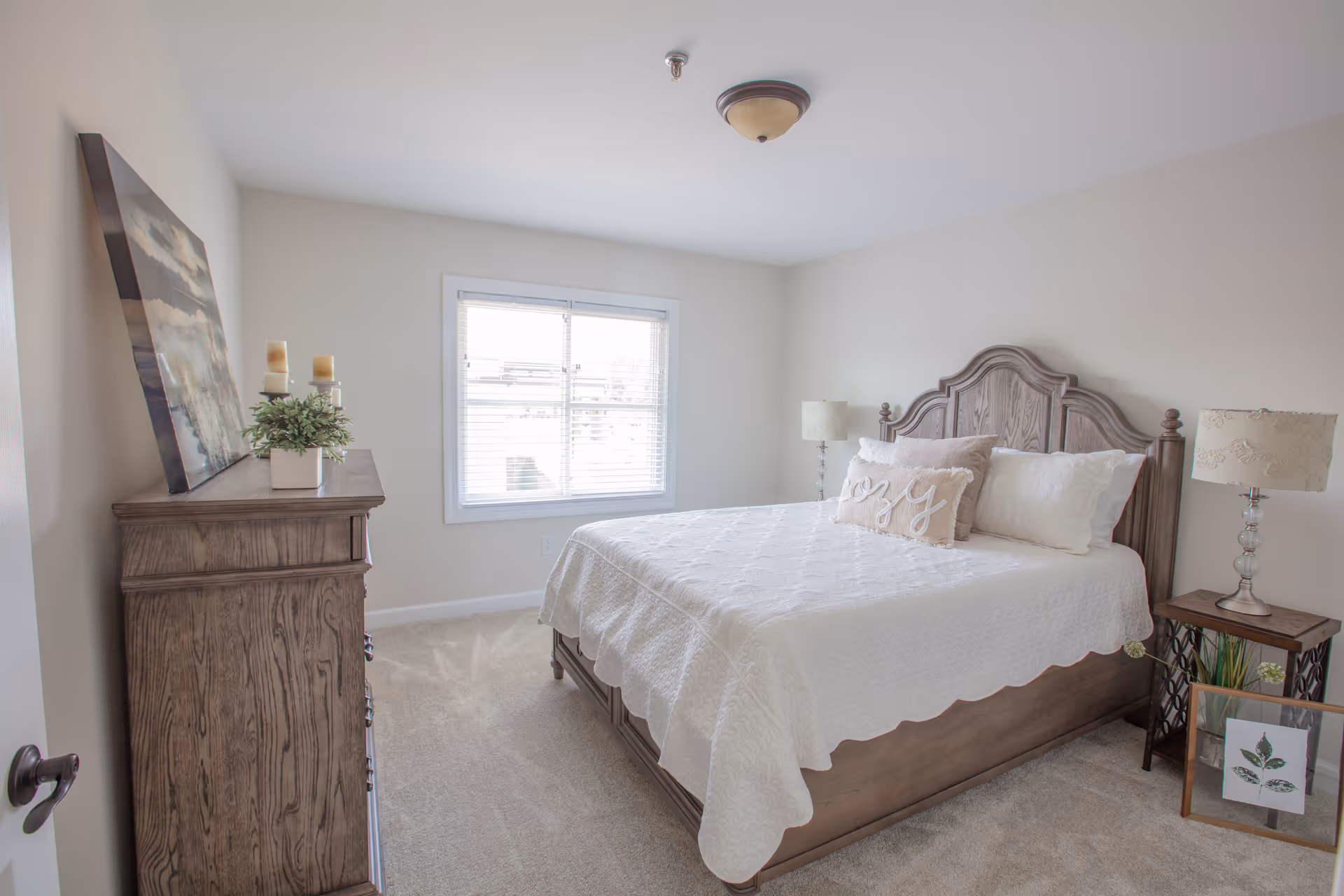 A bright and cozy bedroom with a large wooden bed featuring a white quilt and decorative pillows. There is a wooden dresser with candles and a plant on top, a window with blinds letting in natural light, and two bedside tables with lamps. A framed picture with a leaf design rests on the floor near one bedside table.