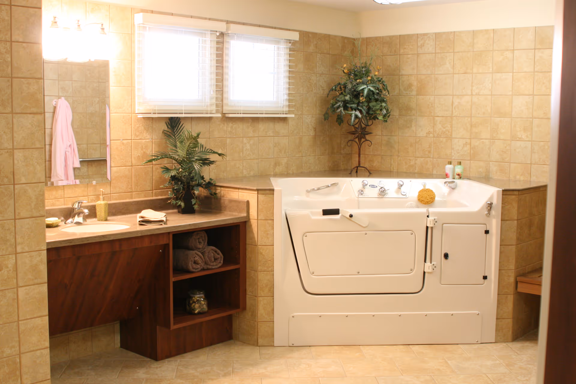A bathroom with beige tiled walls and floor featuring a walk-in bathtub with safety handles and controls, a wooden vanity with a sink, a mirror, folded towels on open shelves, a potted plant on the counter, and a pink robe hanging on the wall. Two small windows with blinds are above the bathtub.