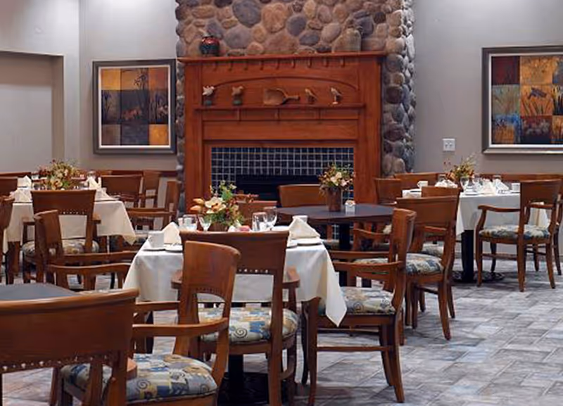 Dining room with multiple wooden tables and chairs, each table covered with white tablecloths and set with glassware and napkins. A stone fireplace with a wooden mantle is centered on the back wall, flanked by two framed abstract paintings. Floral centerpieces decorate the tables.