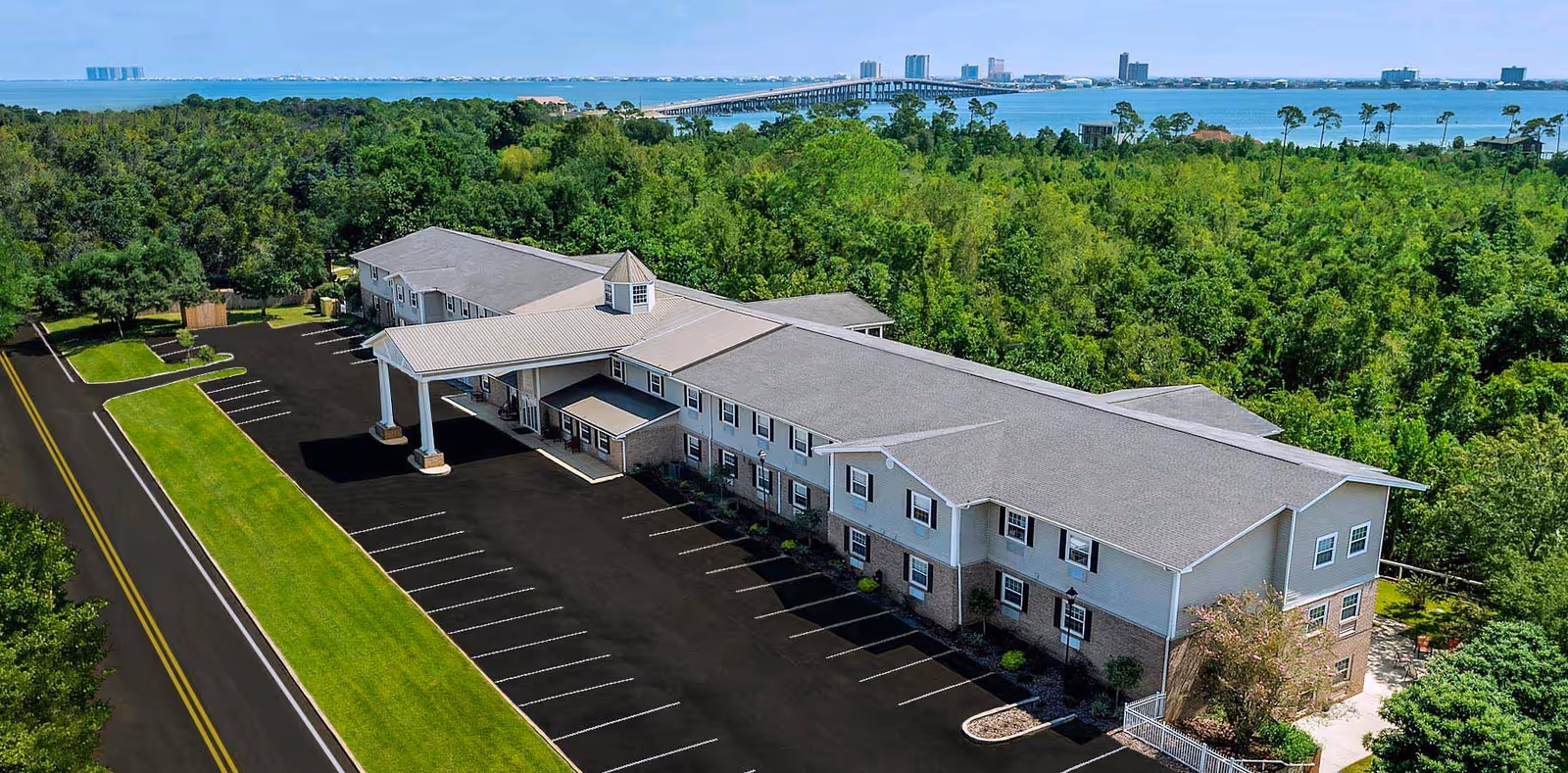 Aerial view of The Camellia of Gulf Breeze senior living facility showing a two-story building with a covered entrance, surrounded by trees and greenery, with a parking lot in front and a body of water in the background.