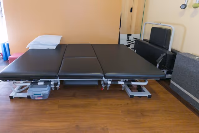 A black padded adjustable therapy table with a pillow in a small room with wood flooring and folded chairs against the wall.