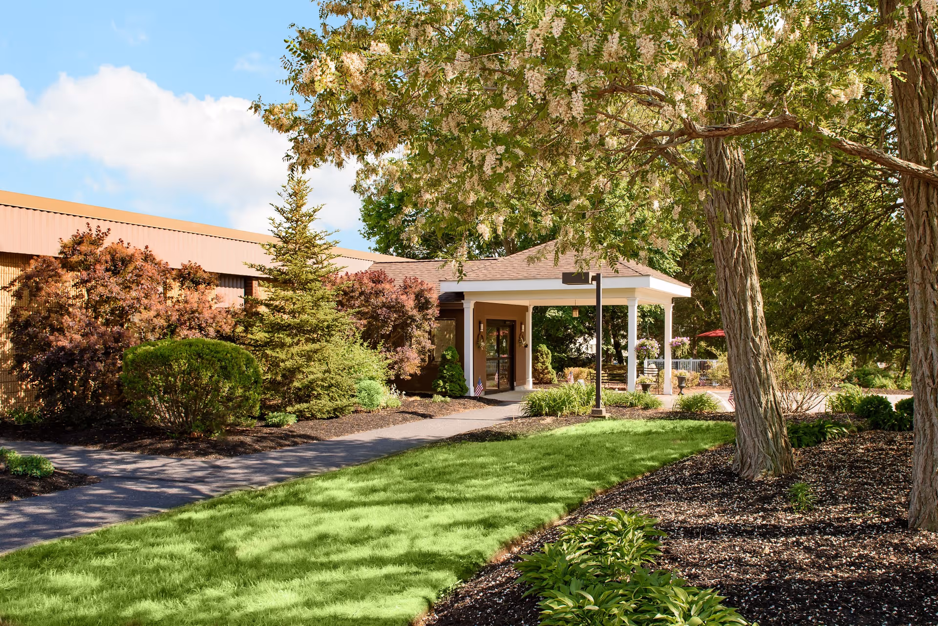 Exterior view of Life Care Center Scituate showing a building entrance with a covered porch, surrounded by well-maintained landscaping including green grass, trees, and shrubs under a partly cloudy blue sky.