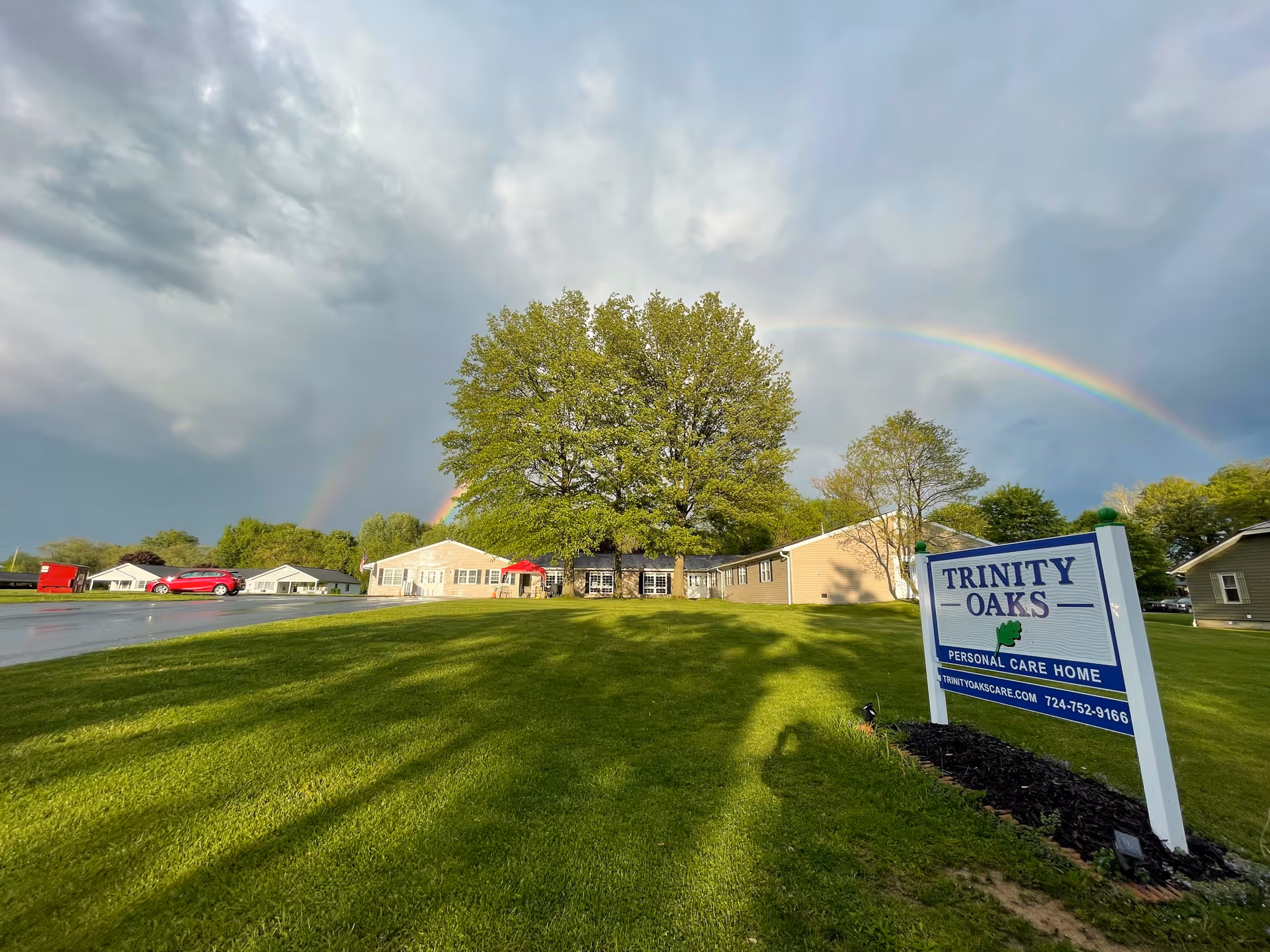Front lawn and sign for Trinity Oaks Personal Care Home with buildings, large trees and a rainbow overhead.