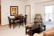A cozy living and dining area in a senior living facility with a dark wooden dining table and four chairs, a framed artwork on the wall, a comfortable armchair, a coffee table with two glasses and a magazine, and a sliding glass door leading to a balcony.