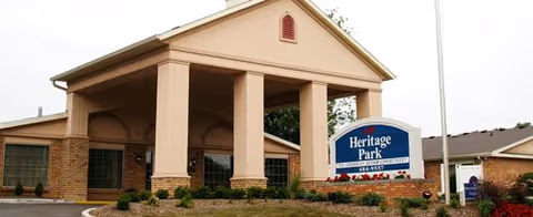 Exterior view of Heritage Park facility showing a covered entrance with large columns, brick walls, and a sign with the facility name in front of the building.