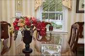 A glossy dining table with a floral centerpiece, wooden chairs, and a window with curtains in the background.