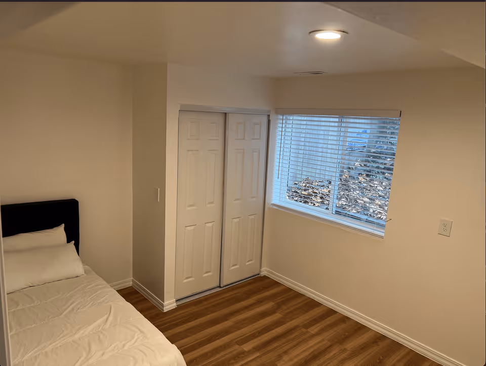 A small bedroom with a single bed against the left wall, white bedding, a window with blinds on the right wall, and a closed closet with double doors next to the window. The room has light-colored walls and wood flooring.