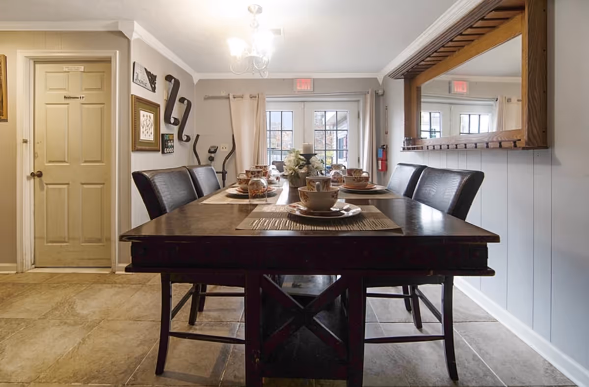 A dining room with a dark wooden table set for six people with matching chairs. The table has placemats, cups, and a centerpiece. There is a large mirror on the right wall, a door labeled 'Bedroom #7' on the left, and a glass door with curtains at the back letting in natural light.