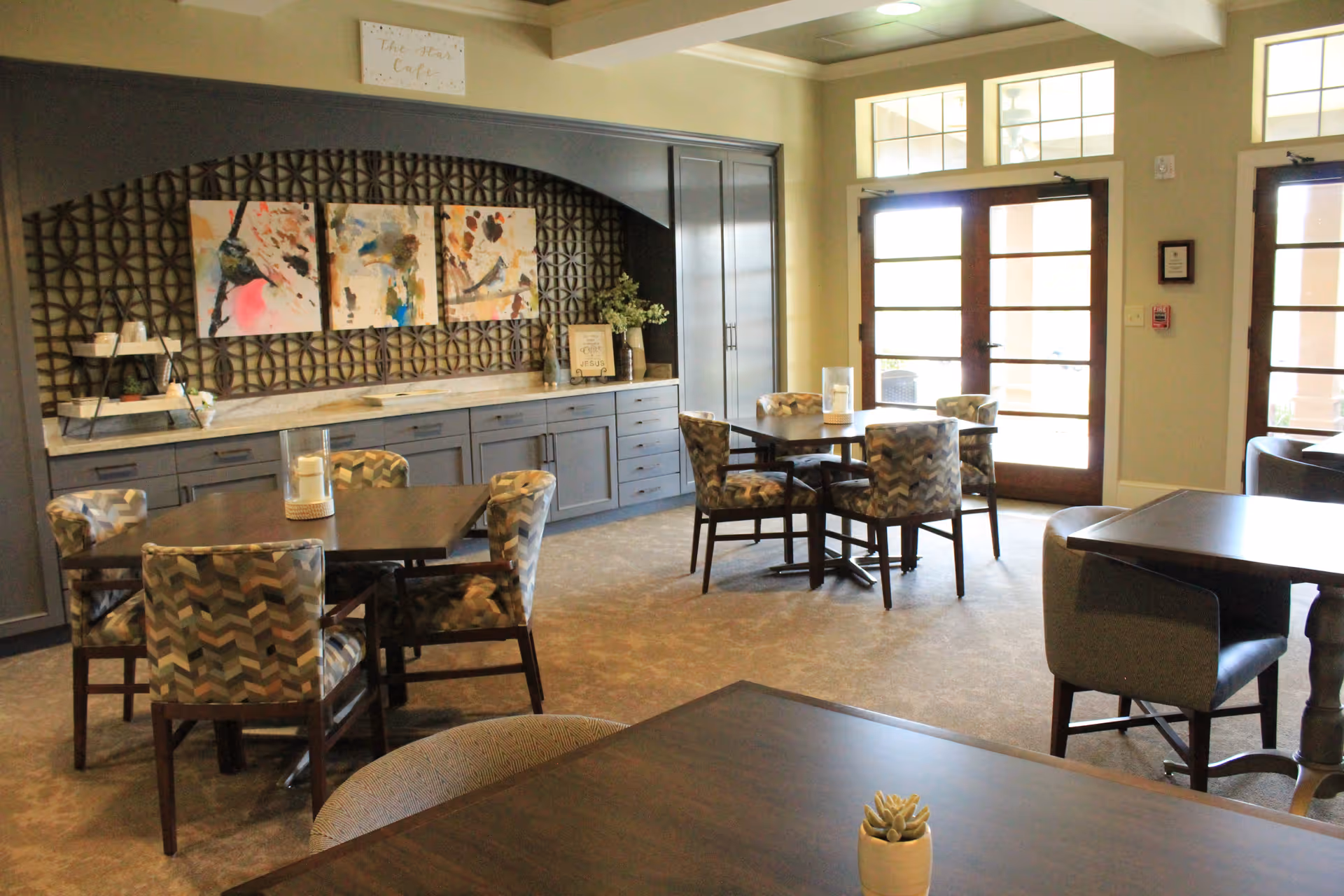 A bright and inviting common area with several tables and patterned chairs arranged for seating. The room features large windows and glass doors allowing natural light to enter. A built-in cabinet with decorative artwork and plants is visible along one wall.