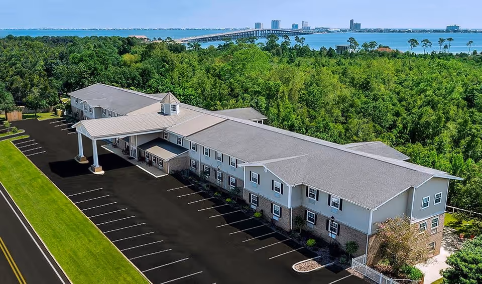 Aerial view of a two-story senior living building with a covered entrance, large parking lot, surrounding trees, and a bay with a bridge in the background.