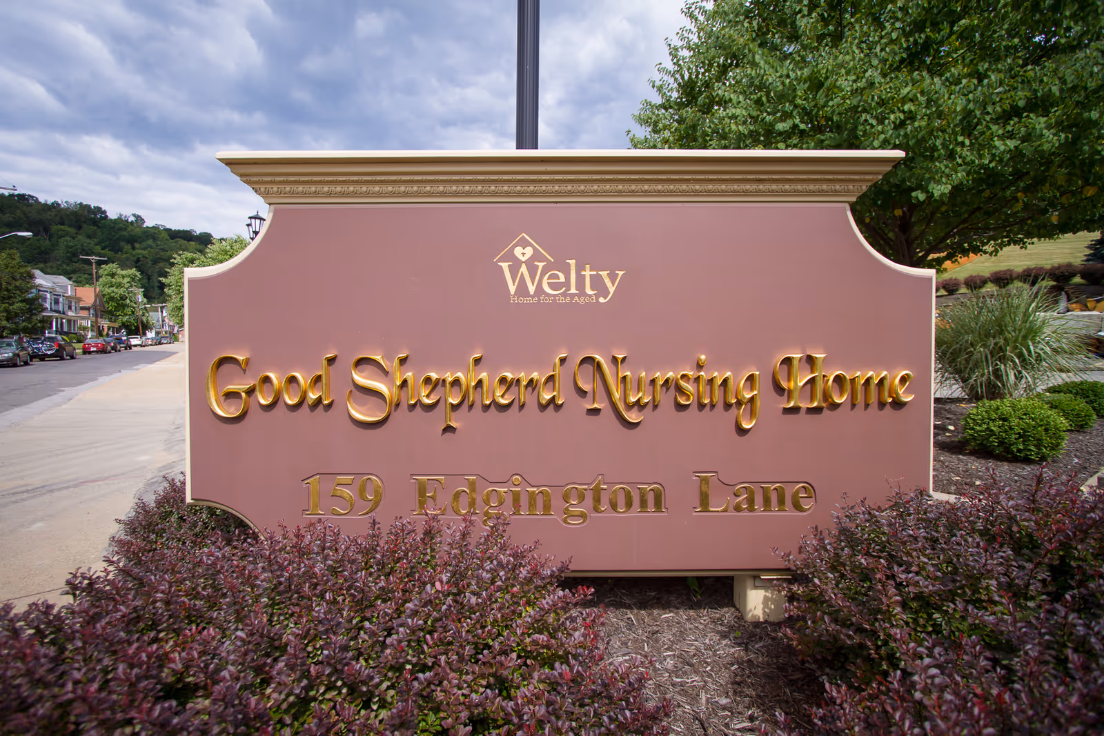 A large outdoor sign for Good Shepherd Nursing Home at 159 Edgington Lane, with the Welty Home for the Aged logo above it. The sign is surrounded by bushes and trees, with a street and houses visible in the background under a cloudy sky.