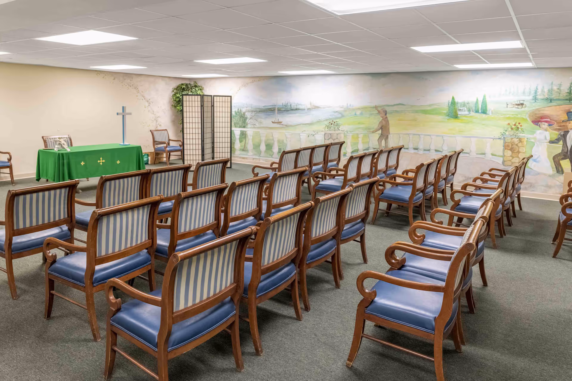 Rows of wooden chairs facing a small altar with a cross and green cloth in a chapel-like room with a scenic wall mural.