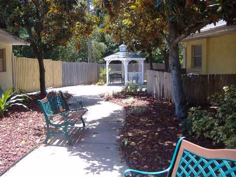 A shaded outdoor walkway with benches on either side, surrounded by trees and landscaping. A white gazebo is visible at the end of the path, with a wooden fence and a yellow building on the right side.