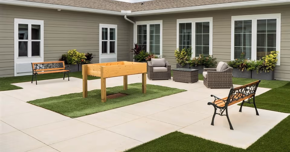 Outdoor patio area at The Canopy at Walden Woods featuring two wooden benches with decorative metal backs, a raised wooden planter box on artificial grass, and a seating area with two cushioned wicker chairs and a matching wicker coffee table. The patio is surrounded by a building with beige siding, white-framed windows, and window boxes with green plants.