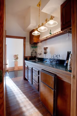 A narrow kitchen area with wooden cabinets and drawers, a dark countertop, and three hanging pendant lights. The kitchen has a small wine rack, decorative items, and a window letting in natural light. The floor is wooden, and there is an open doorway leading to another room with a decorative vase on a pedestal.
