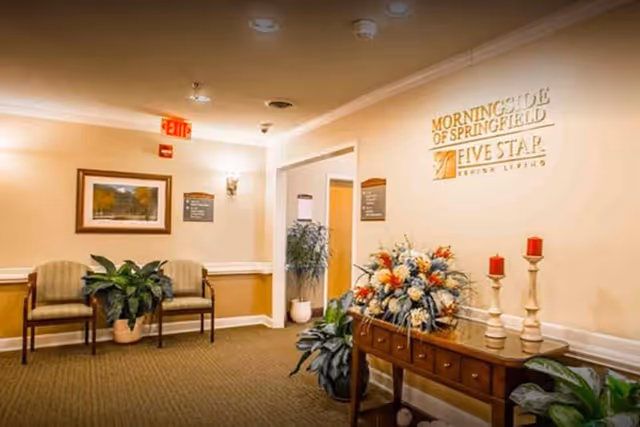 A warmly lit senior living facility interior with a waiting area of chairs and plants, a console table topped with flowers and candles, and a wall sign reading 'Morningside of Springfield'.