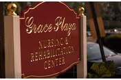 A red and gold sign for Grace Plaza Nursing & Rehabilitation Center mounted on white posts with decorative gold finials, with a blurred background showing part of a car and some greenery.