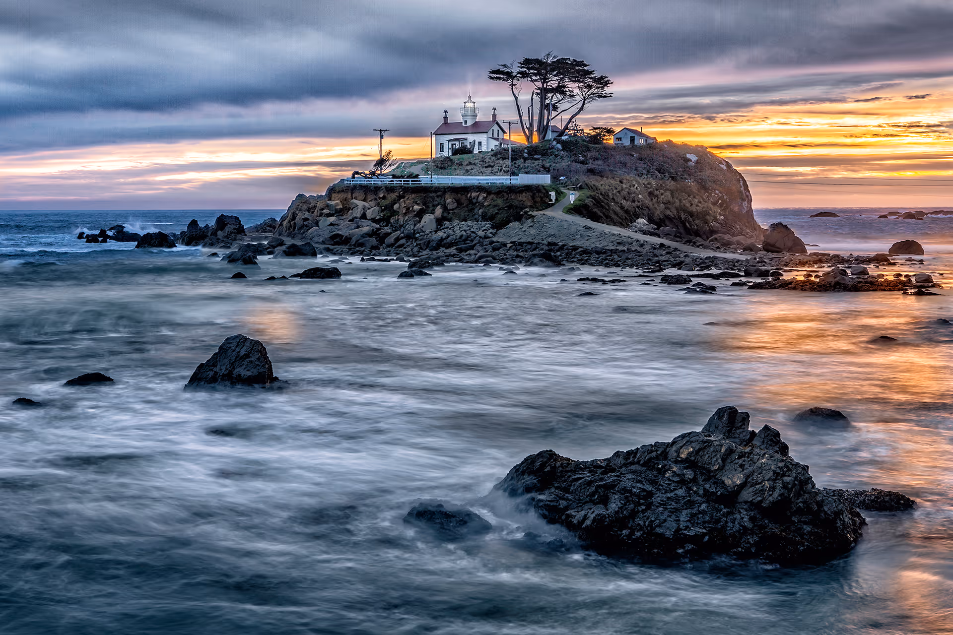 A rocky shoreline and small island topped by a house and lighthouse with trees, lit by a colorful sunset over the ocean.