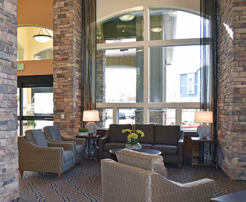 A cozy seating area inside a building with a large window letting in natural light. The area features a dark sofa, two patterned armchairs, a round coffee table with a flower arrangement, two side tables with lamps, and stone pillars on either side of the window.
