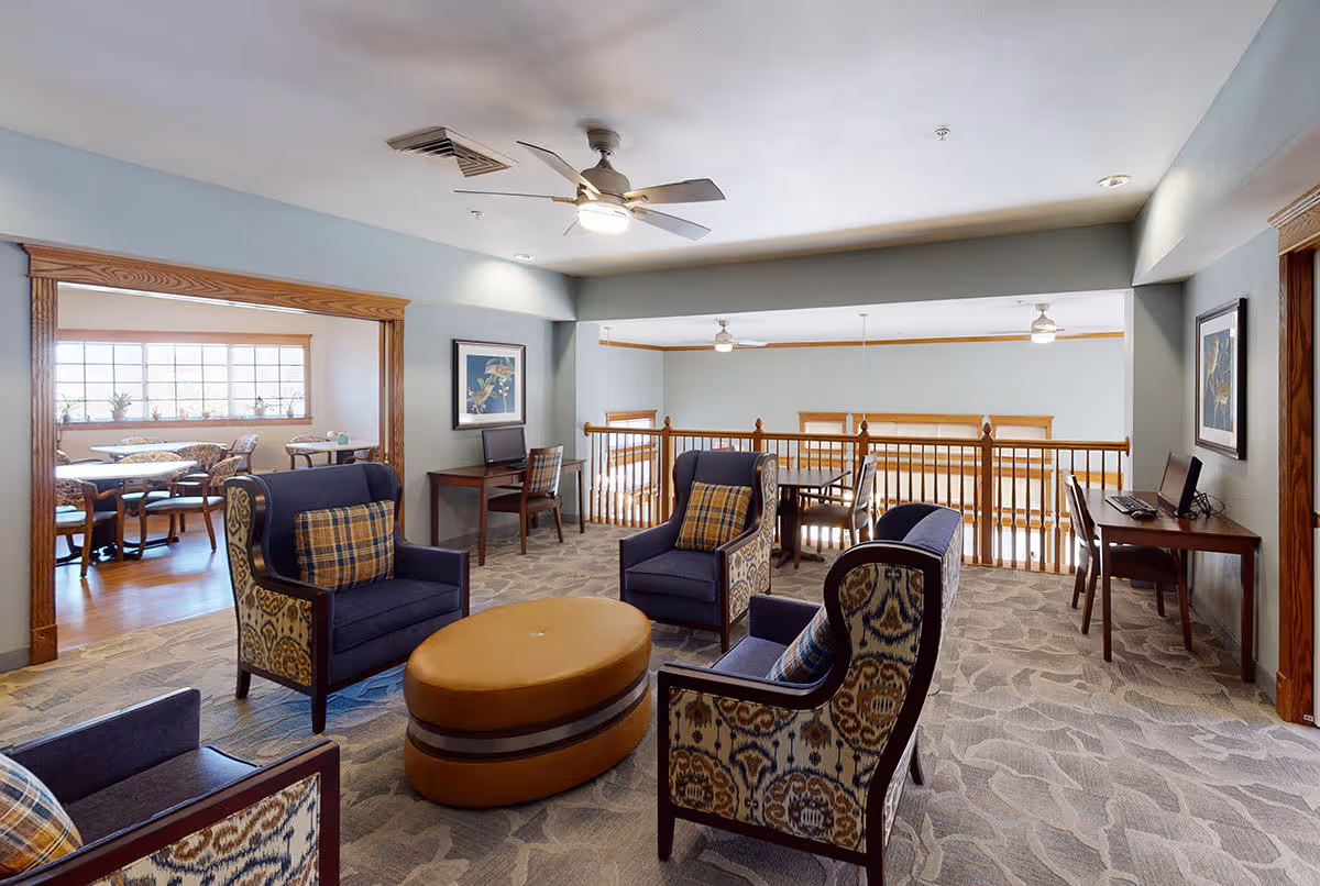 A cozy senior living facility common area with four upholstered armchairs arranged around an oval ottoman. The room has carpeted flooring and light blue walls with wooden trim. Two desks with computers and chairs are positioned against the walls. In the background, there is a dining area with tables and chairs near a large window letting in natural light. Ceiling fans and recessed lighting illuminate the space.