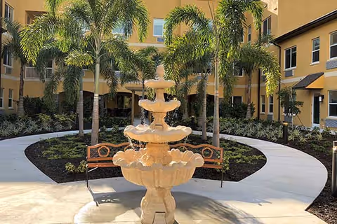 Outdoor courtyard area at Aravilla Sarasota featuring a multi-tiered stone fountain in the center, surrounded by palm trees, landscaped garden beds, and curved concrete walkways. Two wooden benches with metal armrests are placed near the fountain. The courtyard is enclosed by a yellow building with multiple windows.
