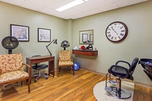 A small salon room with two patterned armchairs, a wooden desk with a black swivel chair, two hair drying stations, a large wall clock, a mirror, and a black salon chair next to a hair washing sink. The floor is wooden and the walls are painted light green with two framed pictures.