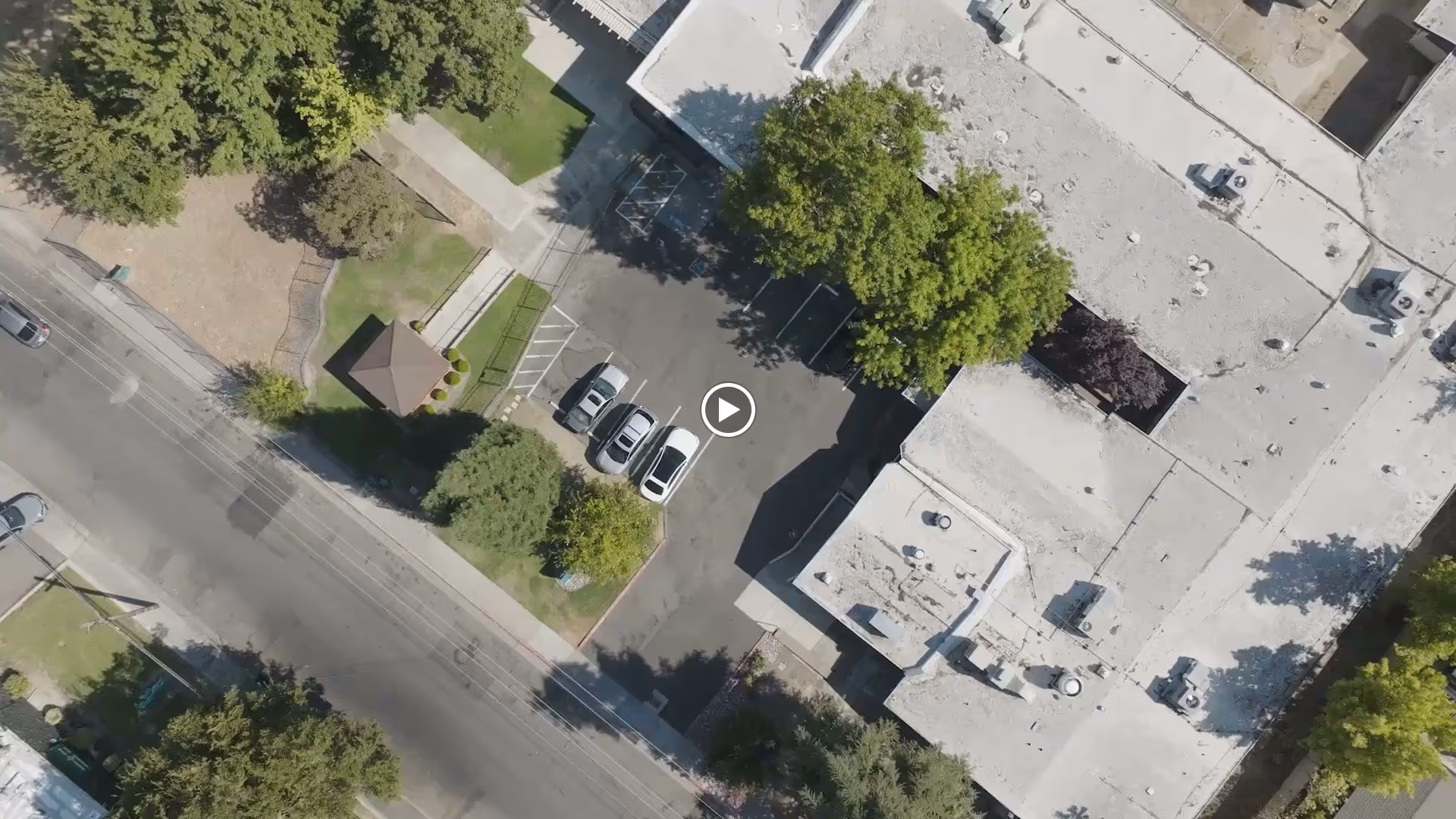 Aerial view of a building and adjoining parking lot with several parked cars, nearby trees, and a small gazebo by the street.
