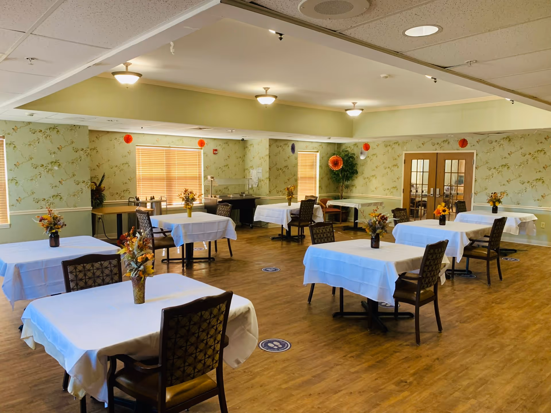 Spacious dining room with multiple tables covered in white tablecloths, chairs, floral centerpieces, and wood flooring.