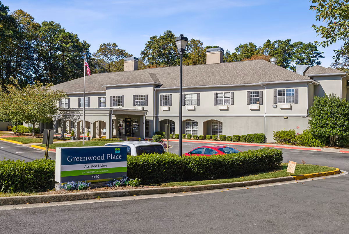 Two-story beige assisted living building with arched entry, landscaped hedges, parked cars, and a 'Greenwood Place' sign in the foreground.