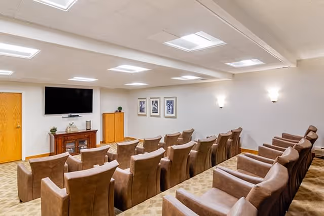 A small theater room with two rows of brown leather recliner chairs facing a flat-screen TV mounted on a white wall. Below the TV is a wooden cabinet with decorative items. The room has beige carpet, white walls, and ceiling lights.