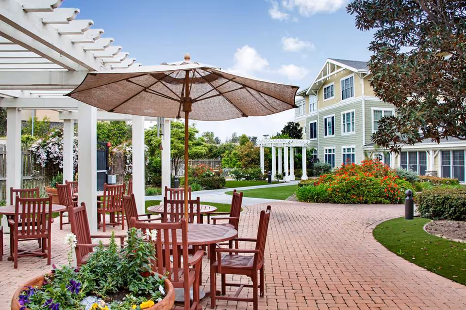 Outdoor courtyard with wooden tables and chairs under a large umbrella, a pergola, landscaped paths and a multi-story residential building.
