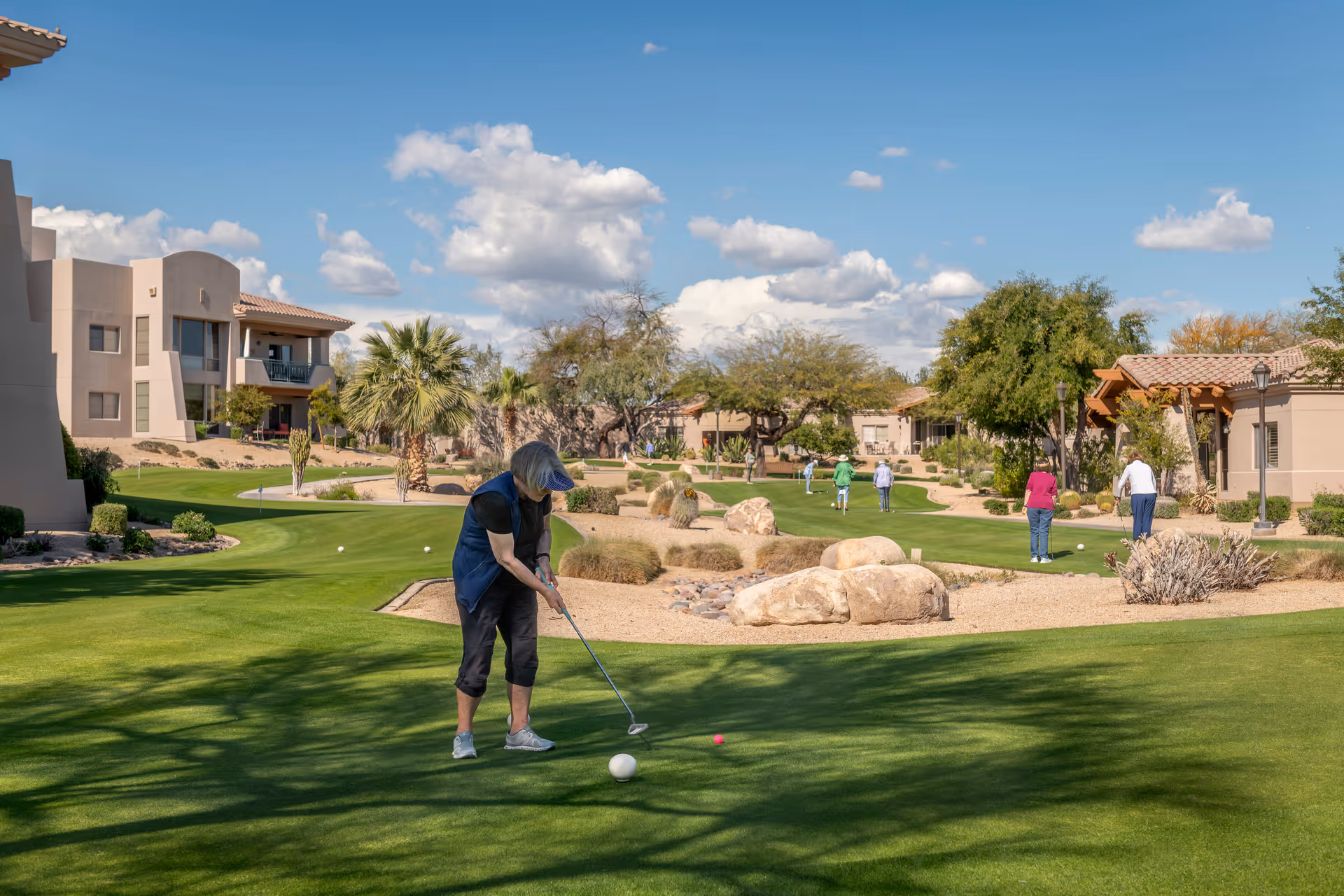 A woman playing golf on a well-maintained putting green surrounded by desert landscaping and residential buildings under a blue sky with scattered clouds. Several other people are also playing golf in the background.