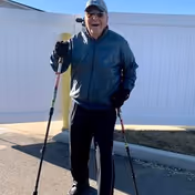 An elderly man wearing a cap, sunglasses, a dark jacket, and gloves is standing outdoors on a paved surface holding two walking poles, smiling towards the camera. There is a white fence and a yellow post in the background.