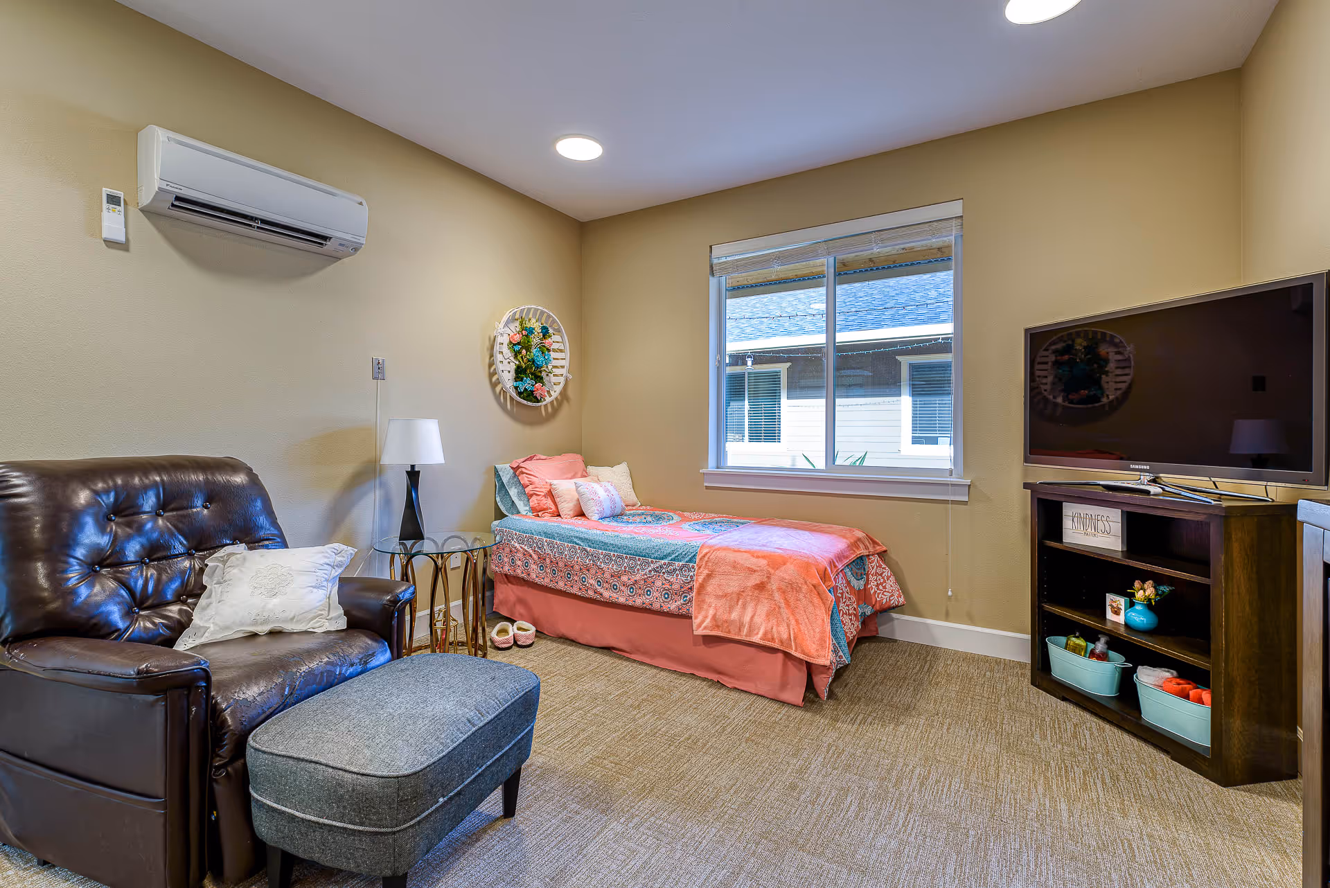 Well-lit memory-care bedroom with a single bed in colorful linens, a leather armchair and ottoman, a TV on a stand, and a window.