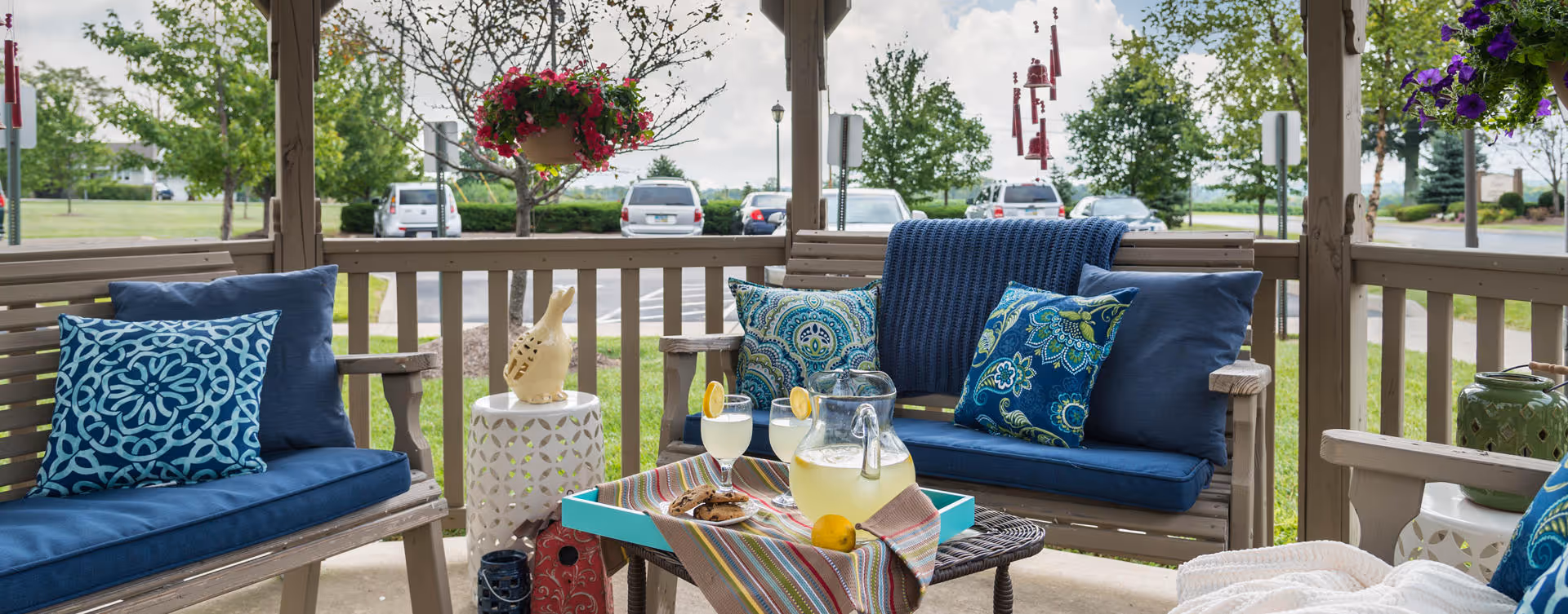 Outdoor covered seating area with wooden benches and blue cushions and pillows. A small table in the center holds a tray with a pitcher of lemonade, two glasses with lemon slices, and a plate of cookies. Hanging flower baskets and wind chimes decorate the space, with a parking lot and trees visible in the background.