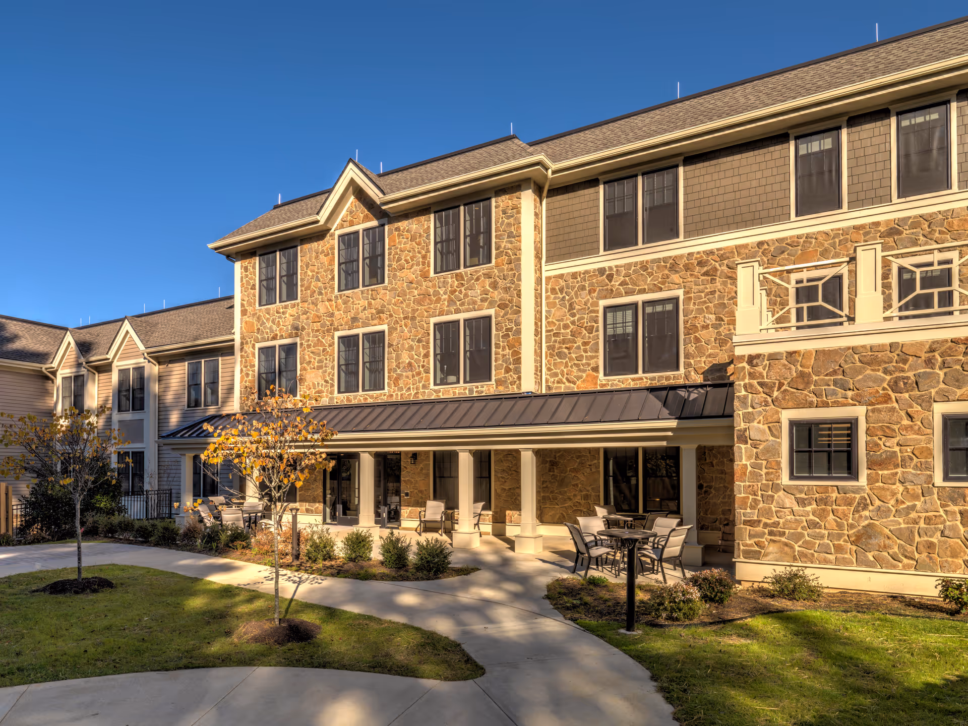 Exterior view of a three-story senior living facility building with stone and siding facade, multiple windows, a covered patio area with outdoor seating, small trees, and landscaped greenery under a clear blue sky.