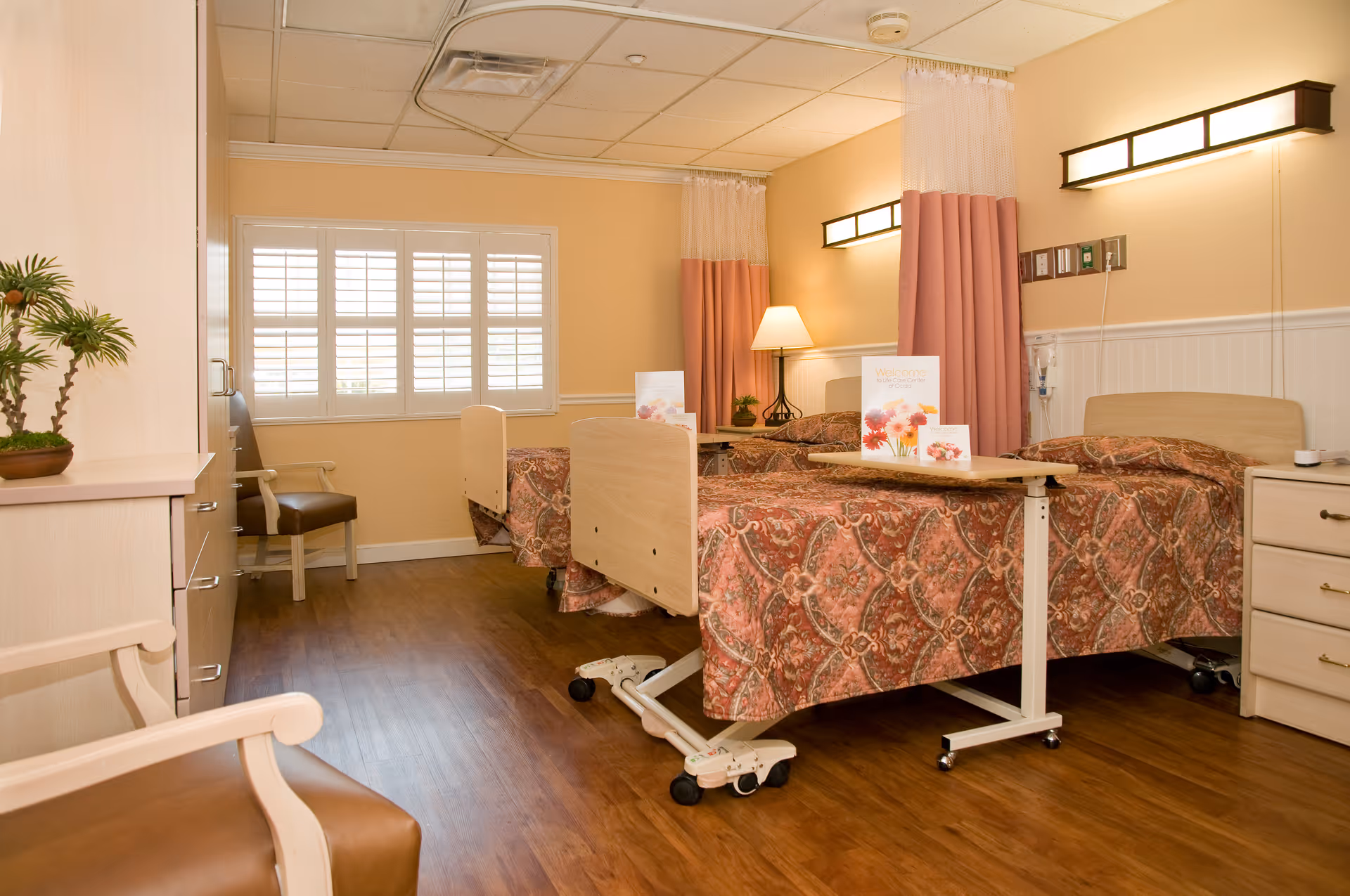 A bright senior-care bedroom with two adjustable beds, bedside tables, chairs, and a window with shutters.