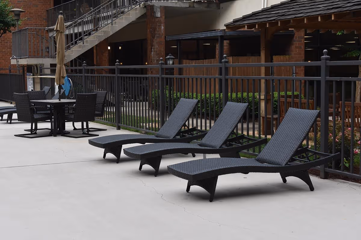 Outdoor patio area with three black woven lounge chairs lined up on a concrete surface next to a black metal fence. In the background, there is a table with four matching chairs and a closed beige umbrella. A brick building with stairs and windows is visible behind the fence.