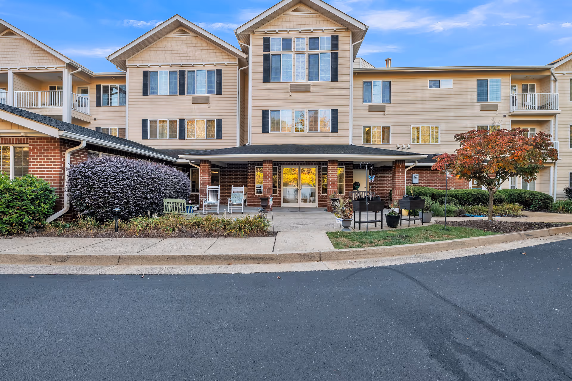 Front entrance of a three-story senior living residence with a covered entry, outdoor seating, and landscaped shrubs.