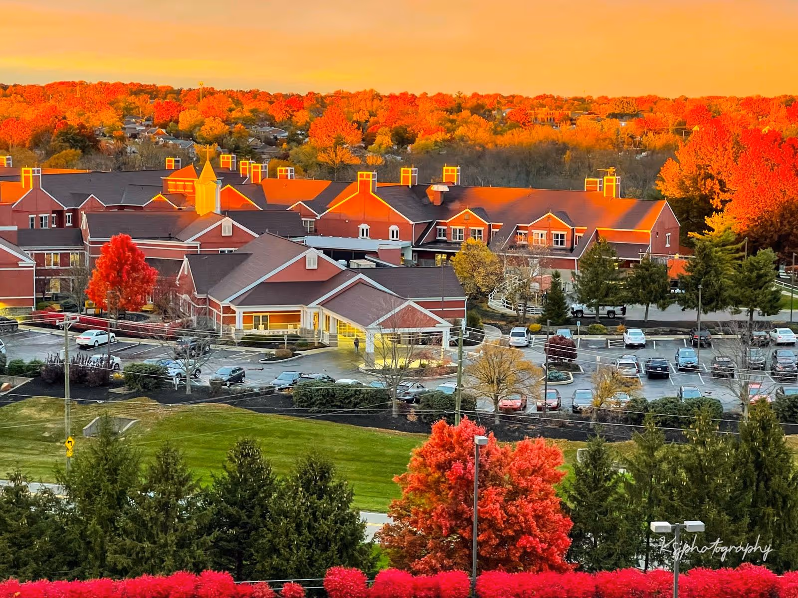 Aerial view of Bayley Senior Care facility surrounded by vibrant autumn foliage with red, orange, and yellow trees. The building has red brick walls and multiple peaked roofs with chimneys. There is a parking lot with several cars and well-maintained green lawns and landscaping around the property.