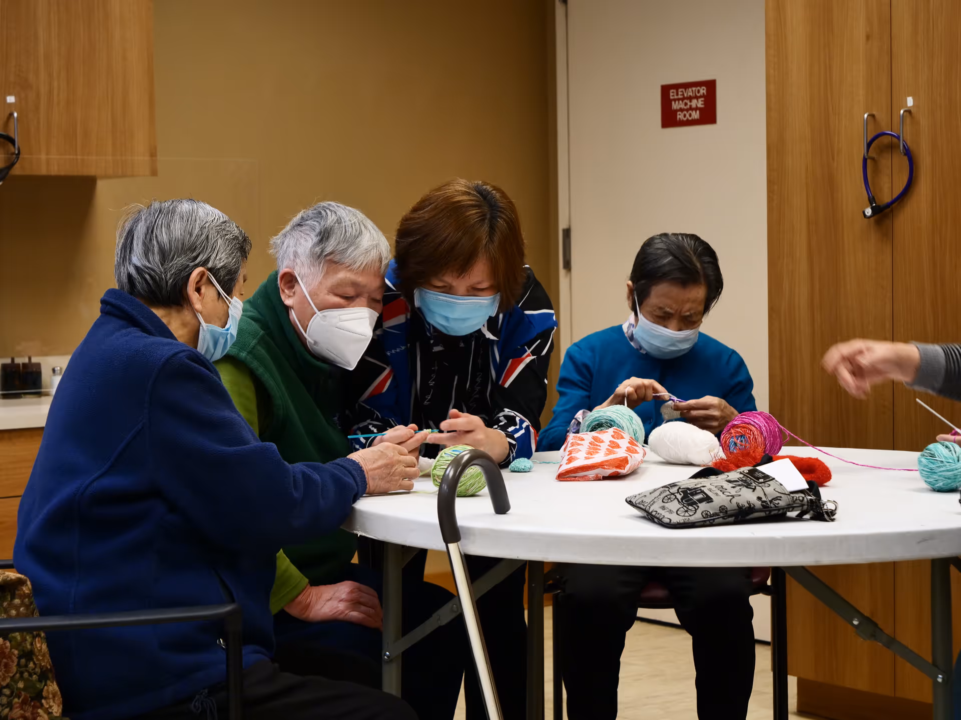 Four elderly individuals wearing face masks sitting around a round table engaged in knitting or crocheting activities with yarn and knitting supplies on the table in a room with wooden cabinets and a door labeled 'Elevator Machine Room'.