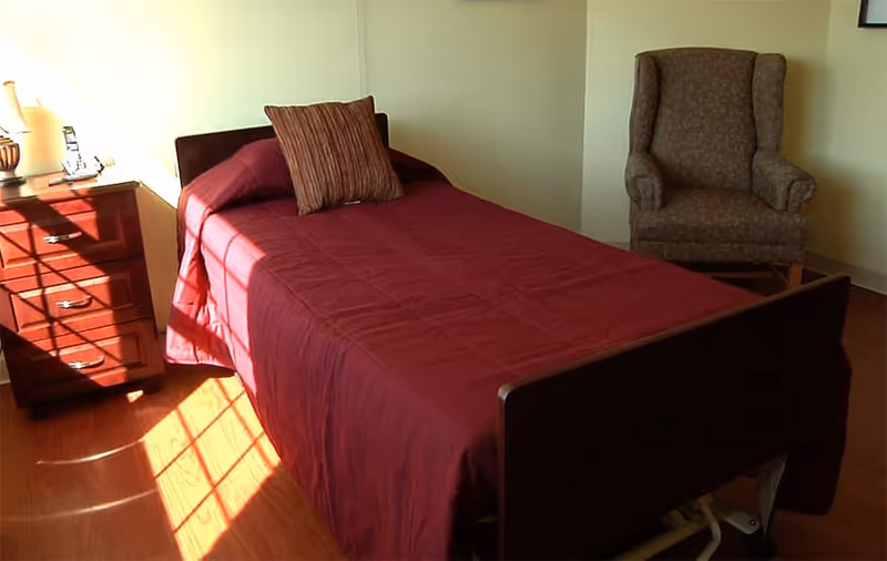 A single bed with a dark red bedspread and a striped pillow in a room with wooden flooring. Next to the bed is a wooden nightstand with drawers, a lamp, and some small items. There is a cushioned armchair with a patterned fabric in the corner of the room. Sunlight is streaming through a window, casting shadows on the floor and bed.