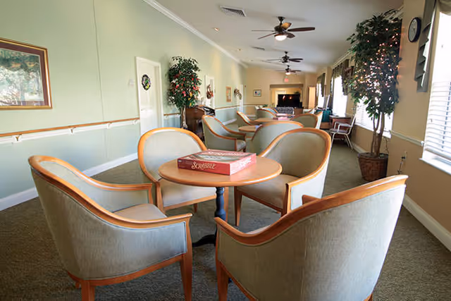 A long, narrow common area with multiple round tables surrounded by cushioned chairs. One table in the foreground has a Scrabble board game on it. The room has light green walls, ceiling fans, potted plants, and windows with blinds on the right side.