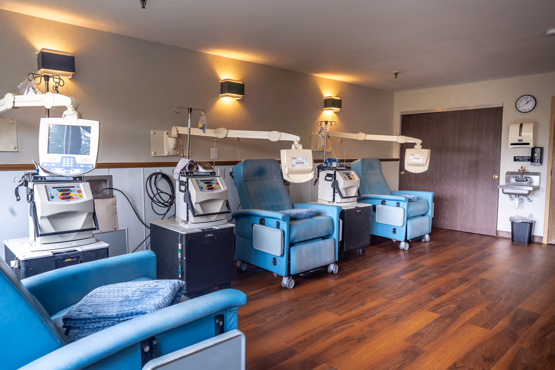 A medical treatment room with four blue reclining chairs, each equipped with attached medical devices and monitors. The room has wooden flooring, soft wall lighting, a clock, a hand washing station with soap dispenser, and a closed wooden door in the background.